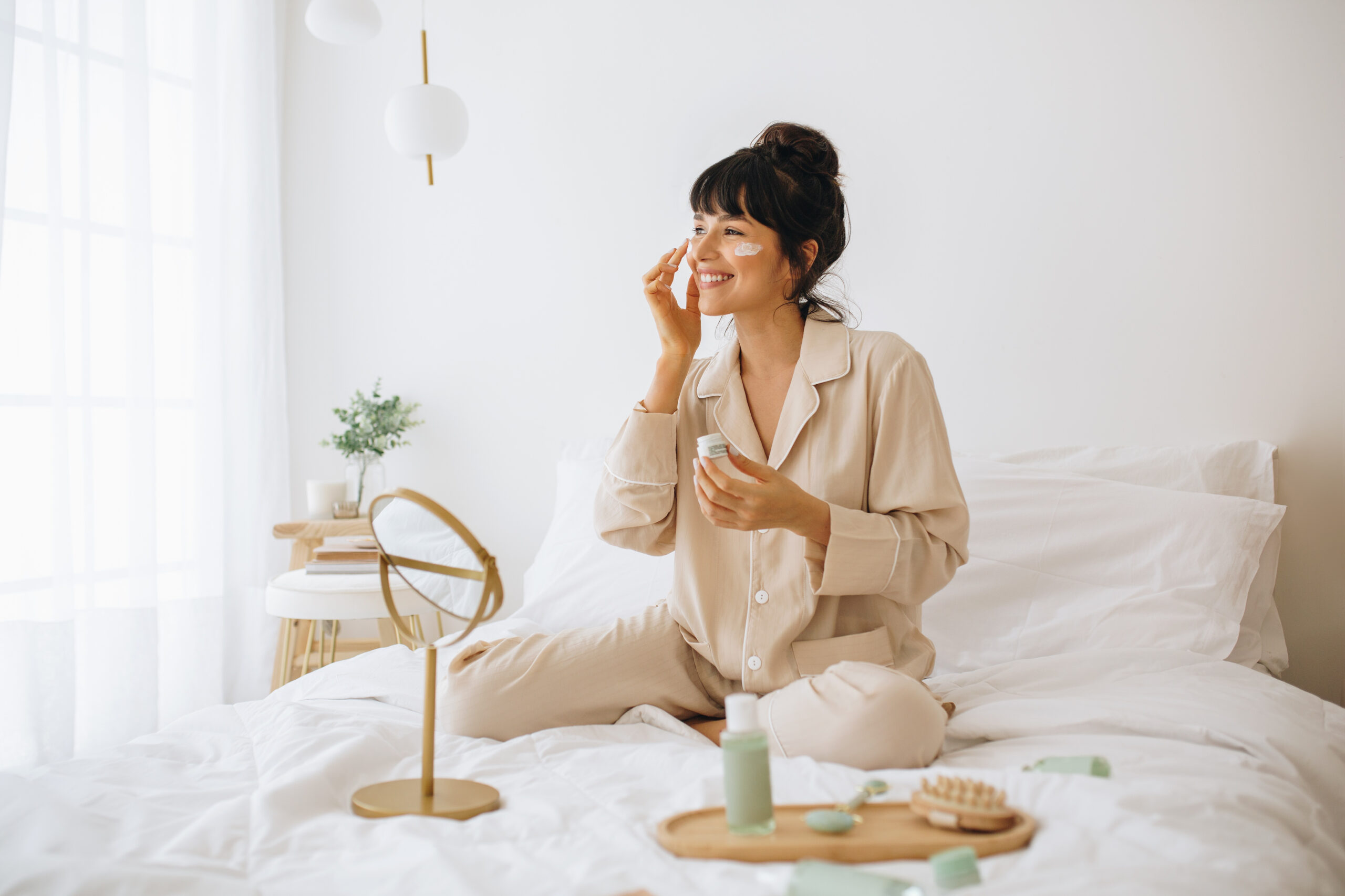 Smiling woman applying face cream sitting on bed Happy woman doing routine skin care at home with beauty products. Woman sitting on bed at home and applying face cream.